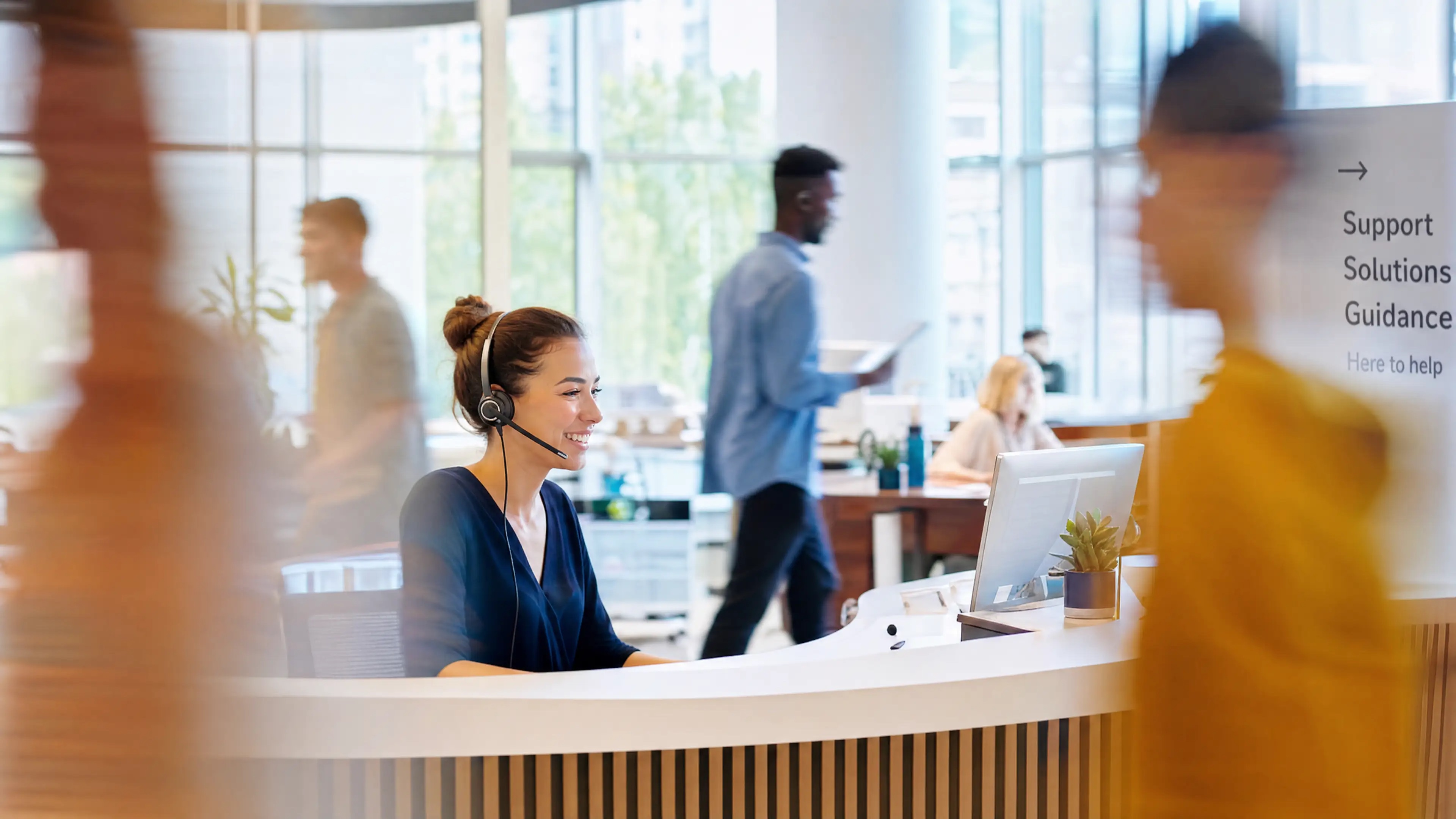 Support agent in a headset smiling at a front desk in a busy office